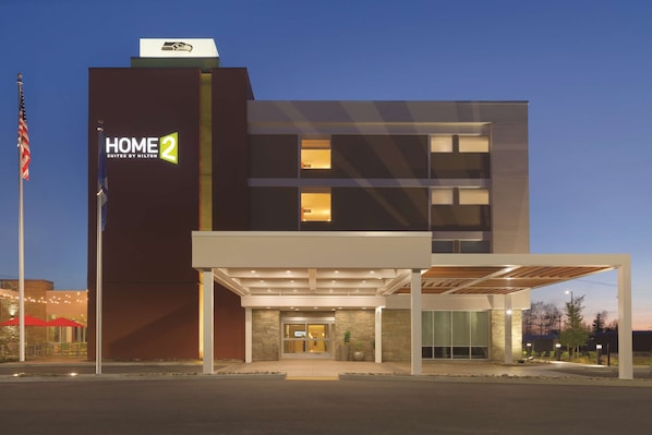 A modern hotel with a prominent "HOME 2 Suites by Hilton" sign, a covered entrance, and a flagpole with an American flag.