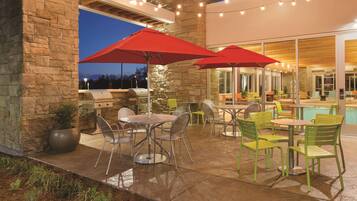 An outdoor patio area with stone walls, metal chairs, tables, red umbrellas, and string lights.