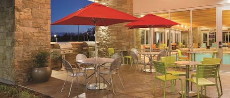 An outdoor patio area with stone walls, metal chairs, tables, red umbrellas, and string lights.