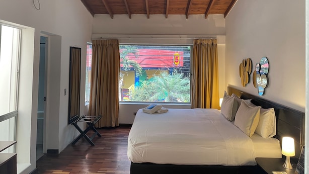 Een hotelkamer met een bed, een bedlamp, een raam met gordijnen, een houten plafond en uitzicht op buiten.