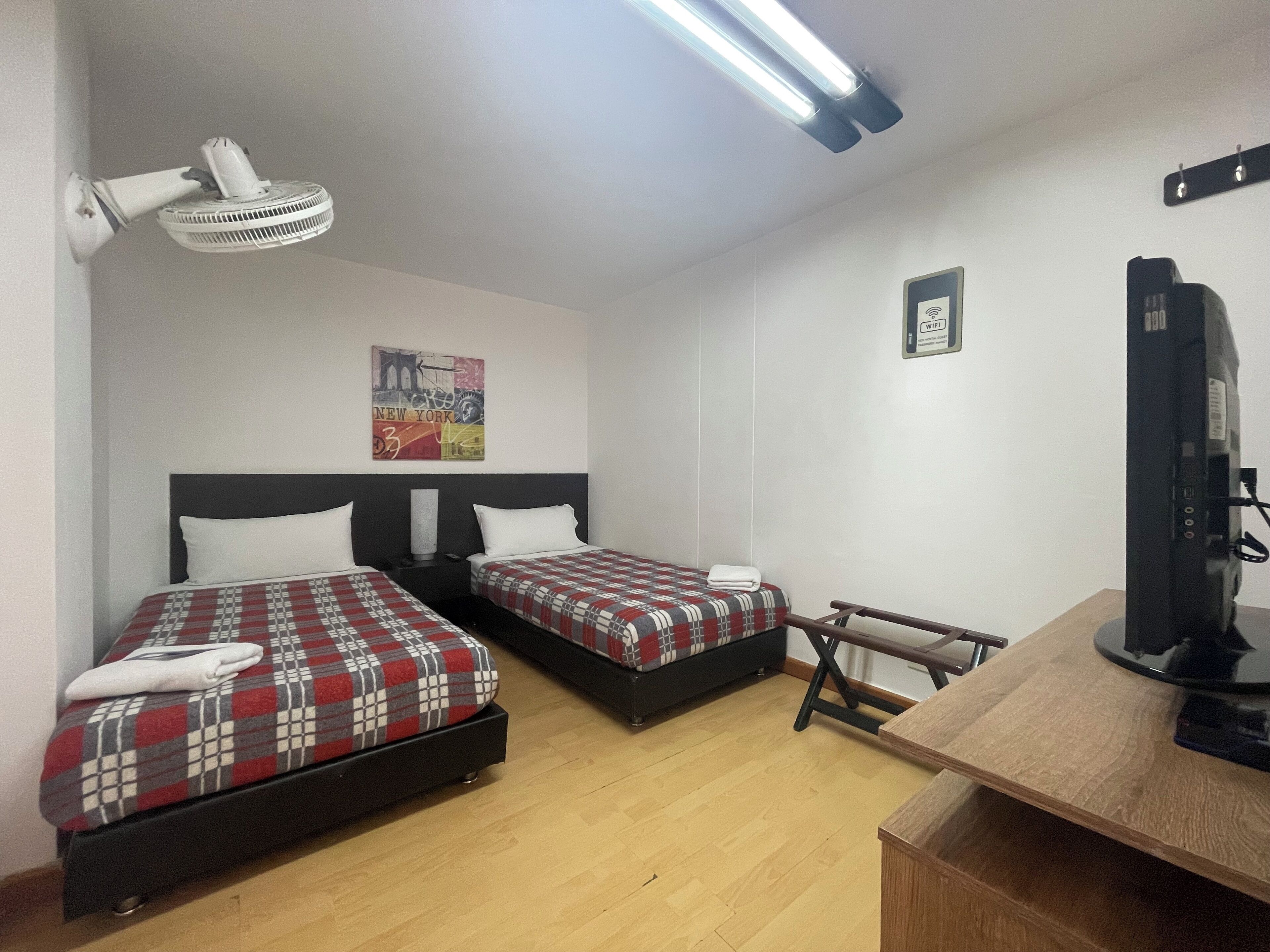 A hotel room with two beds, a desk, a chair, a TV, a ceiling fan, a WiFi sign, and a painting on the wall.