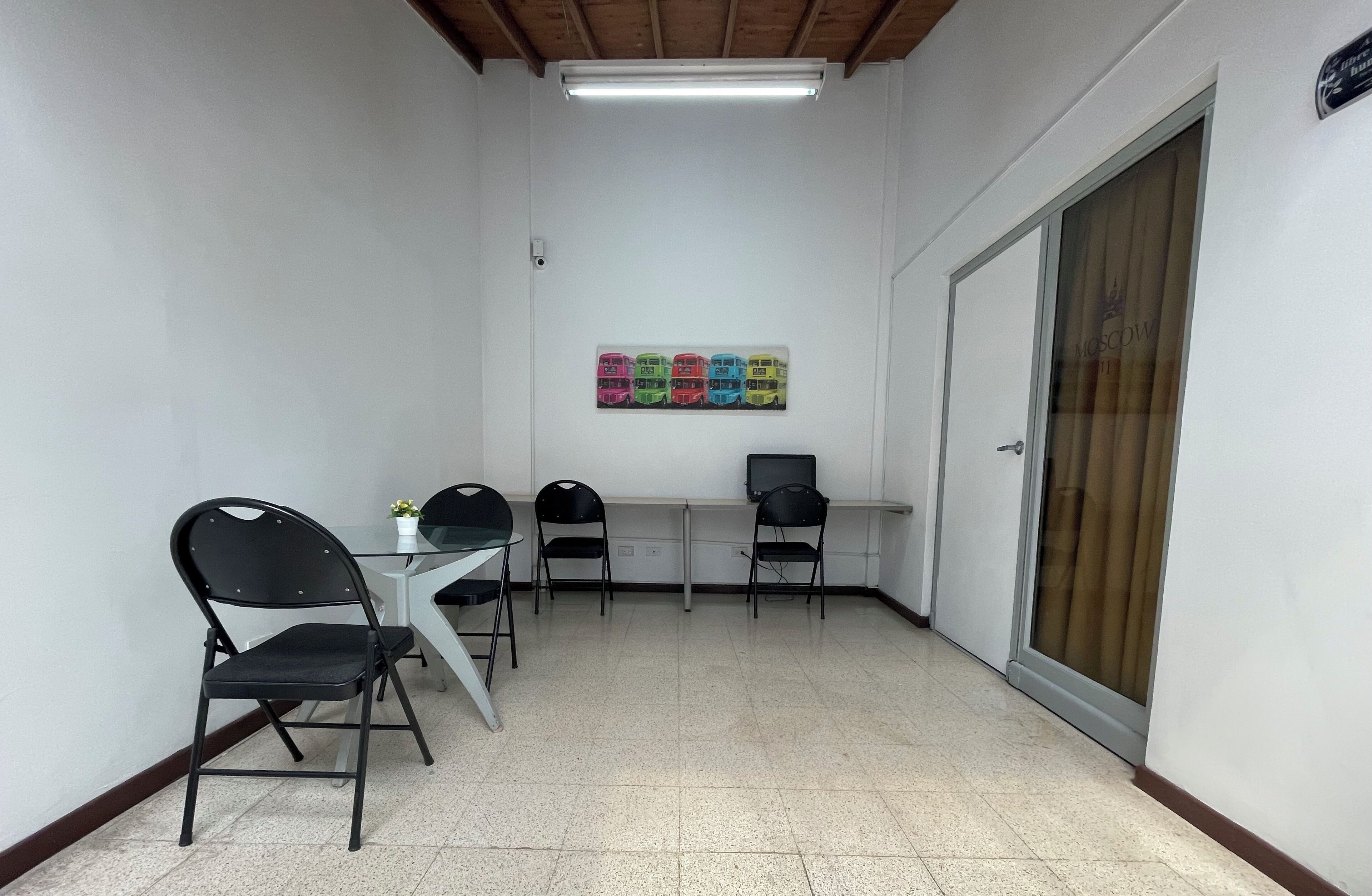 A small, well-lit room with a white table and four black chairs, a flat-screen TV, and a colorful bus artwork on the wall.