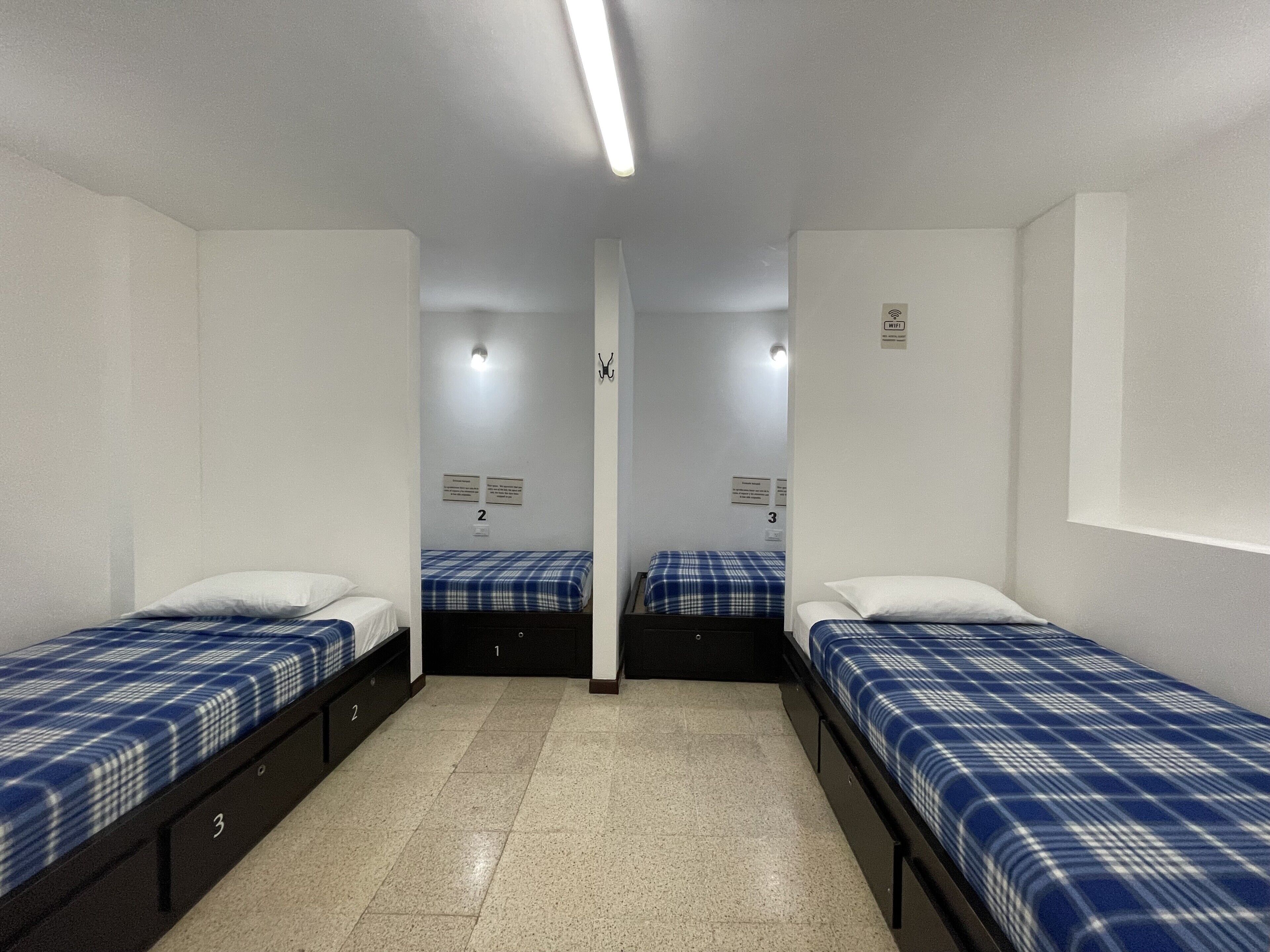Un dormitorio con tres camas individuales, cada una con una colcha azul y blanca a cuadros y almohadas blancas. La habitación tiene paredes blancas, piso de baldosas y una lámpara en el techo. Hay letreros que indican los números de las habitaciones y un símbolo de Wi-Fi en la pared.