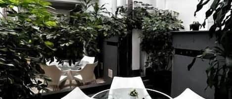 Un patio con una mesa y sillas de vidrio, rodeado de plantas en macetas y un piso de baldosas.