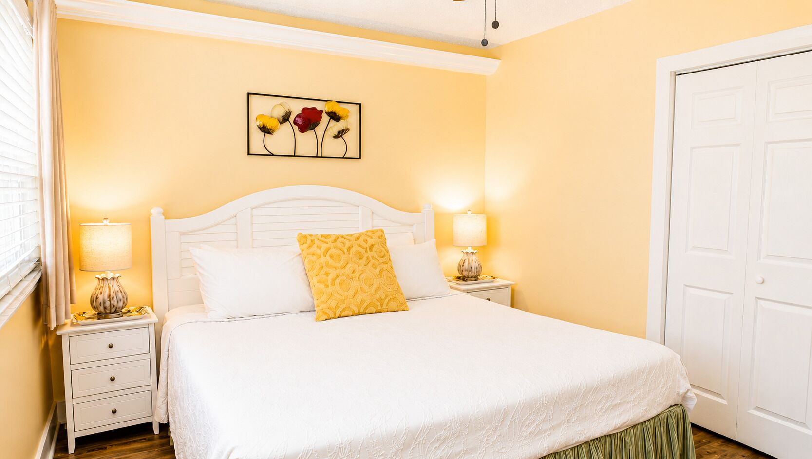 Una cama bien hecha con sábanas blancas y una almohada amarilla, una mesita de noche con lámpara y un cuadro de flores colgado en la pared.