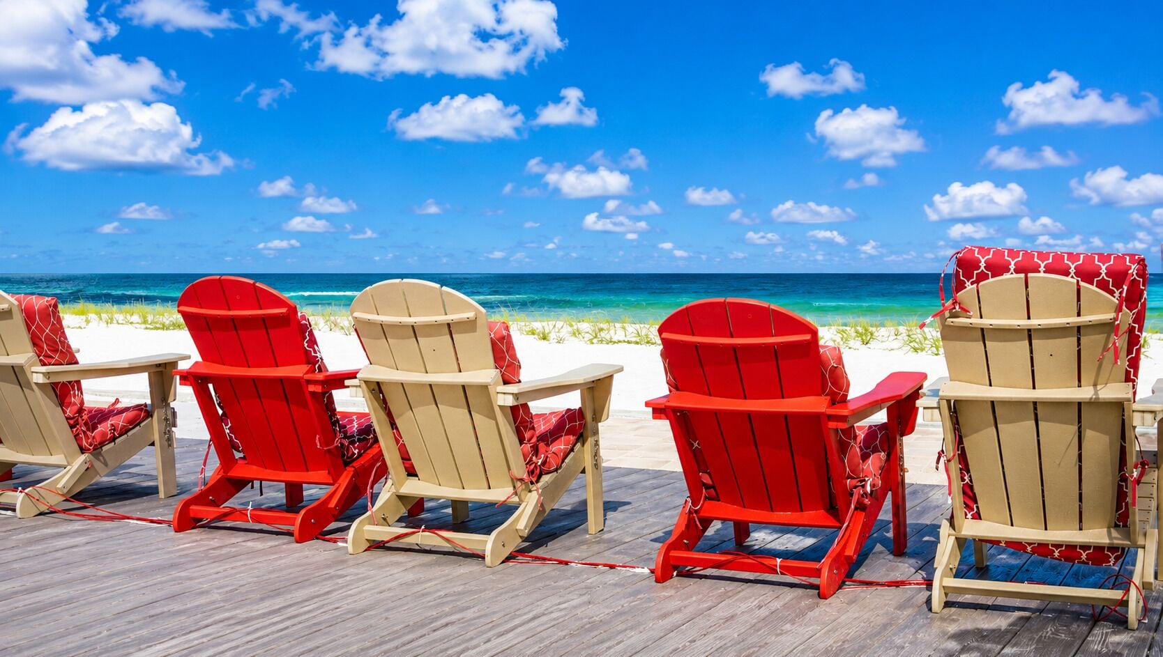 A row of red and beige Adirondack chairs on a wooden deck with a view of the ocean.