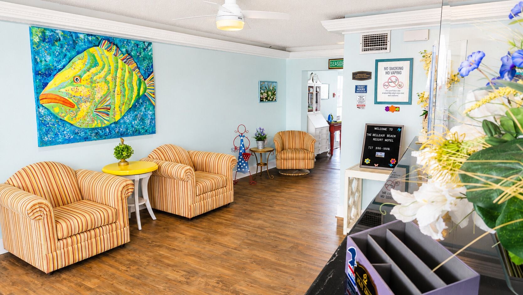 A szálloda előtere csíkos mintájú kanapékkal, egy nagyméretű, halakat ábrázoló festménnyel, egy recepciós pulttal és a Belleair Beach Resort Motel táblájával.