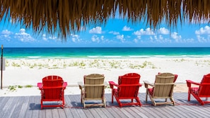 A wooden deck with four red and beige Adirondack chairs facing a beach with clear turquoise water and white sand.