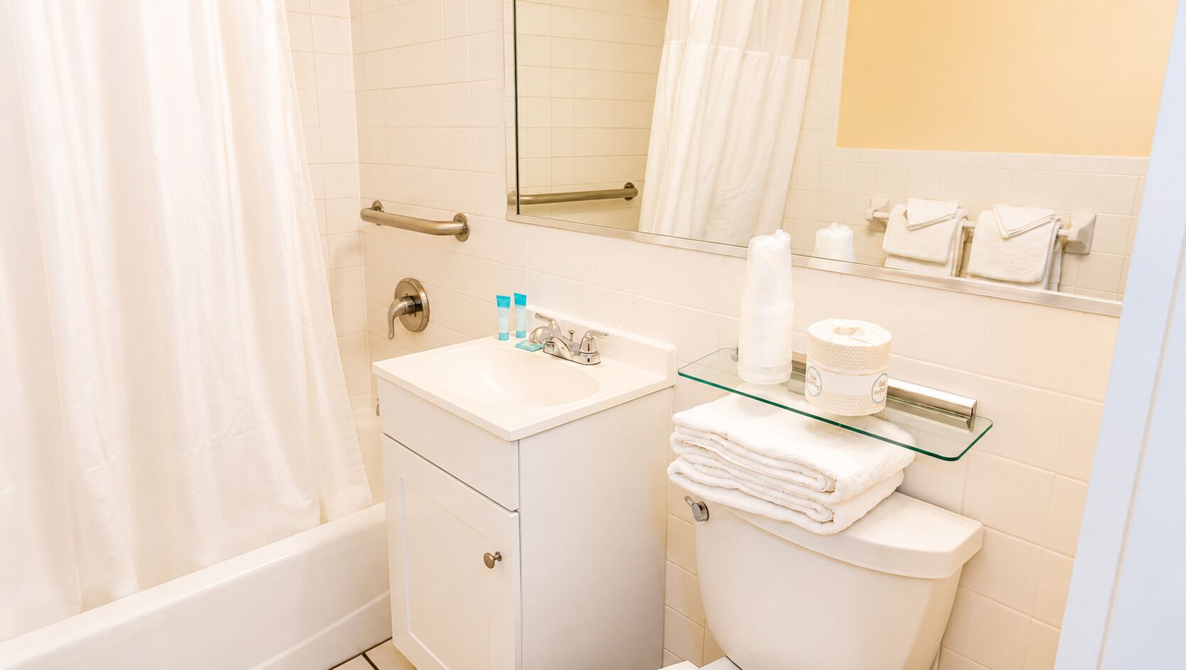 A bathroom with a glass-topped sink, a toilet, a glass shelf with toiletries, and a mirror.