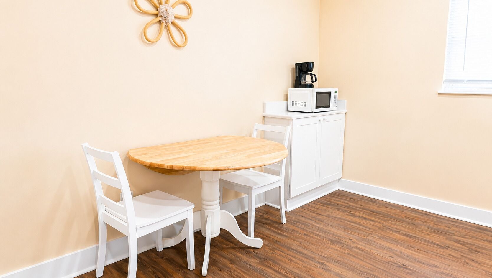 Een eethoek met een ronde houten tafel en witte stoelen, een witte kast met een magnetron en een koffiezetapparaat erbovenop.