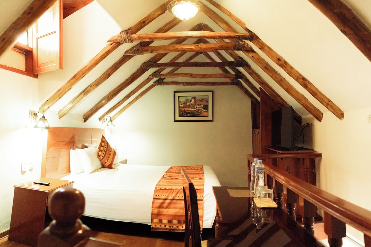 Ein Schlafzimmer mit schrägem Dach, Holzbalken, einem Bett mit Kissen, einem Nachttisch, einer Lampe, einem Gemälde an der Wand und einem Fernseher auf einem Holzboden.