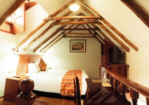 A bedroom with a sloped ceiling, wooden beams, a bed with pillows, a bedside table, a lamp, a painting on the wall, and a TV on a wooden stand. - Hotel Siete Ventanas (Cusco)