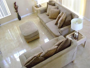 Uma sala de estar com um sofá seccional bege, um banquinho, uma mesa lateral com abajur e uma escultura fixada na parede.