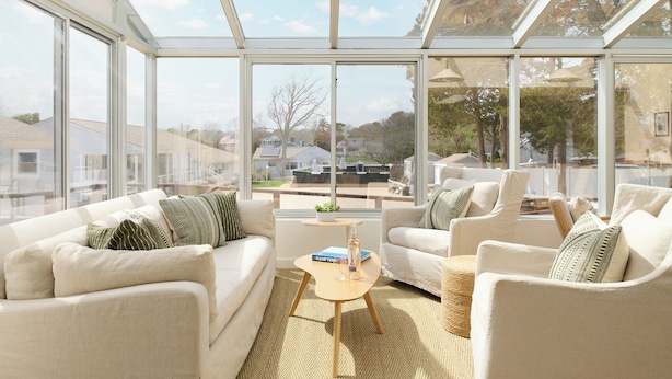 A conservatory with a sofa, chairs, a small table, and a view of houses. - The Penny Lane (Hampton Bays)