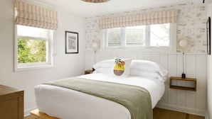 A bedroom with a bed, two bedside tables, a window with curtains, a wall-mounted lamp, and a framed picture. - The Penny Lane (Hampton Bays)