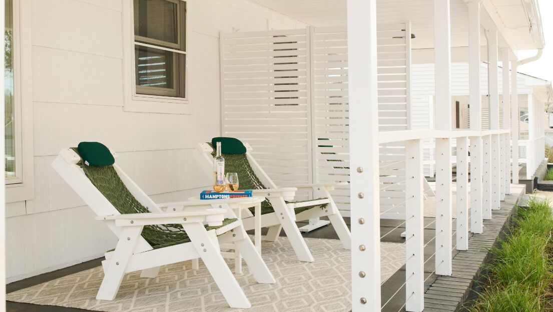 A covered porch with two white lounge chairs, a small table with a bottle and glass, and a book titled 'HAMPTONS.'