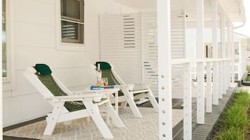 A covered porch with two white lounge chairs, a small table with a bottle and glass, and a book titled 'HAMPTONS.'