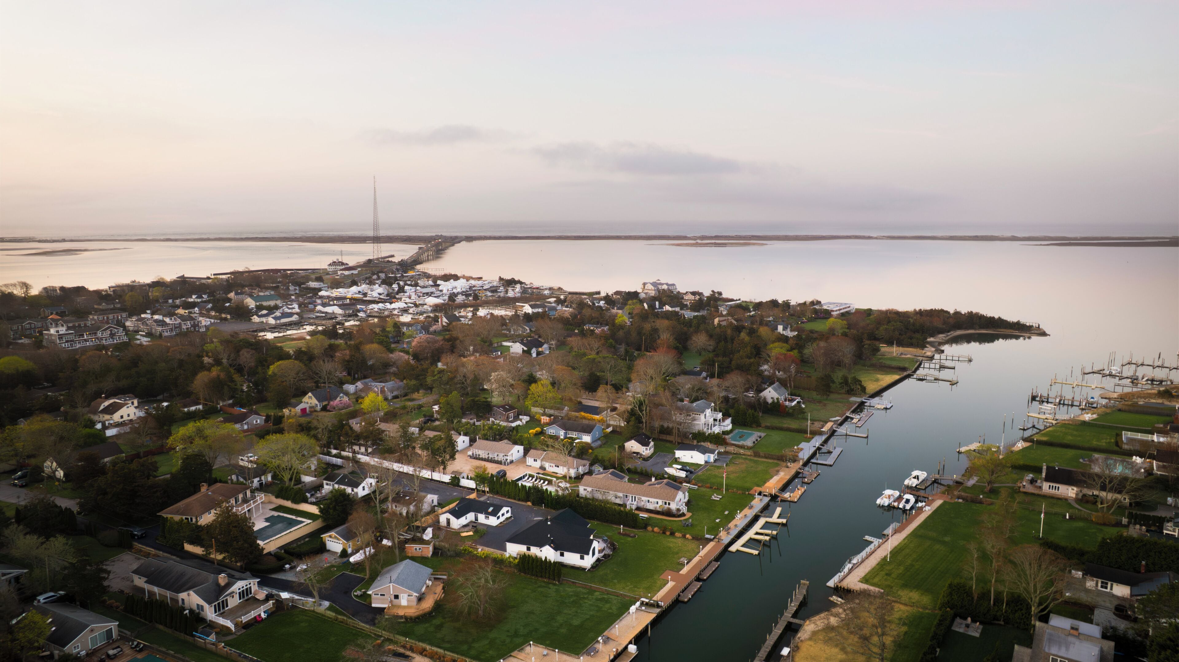 An aerial view of a coastal residential area with houses, a marina, and a pier extending into the water.