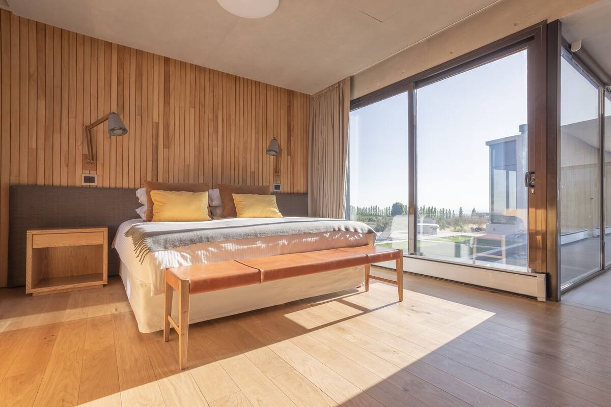 A modern bedroom with a wooden panel wall, a large bed with a bench, bedside lamps, and a sliding glass door leading to a balcony.