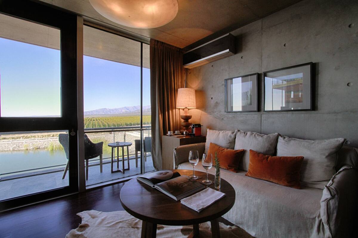 A modern hotel room with a large window offering a view of a lake and mountains. There is a sofa with cushions, a small table with a book and glasses, and a lamp on a side table.