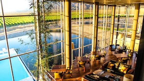 A spacious indoor area with large windows overlooking a river and fields, furnished with sofas, tables, and chairs. - Casa de Uco Vineyards & Wine Hotel (Tunuyan)