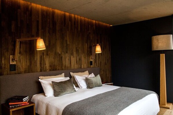 Un dormitorio con cabecera de madera, una cama con varias almohadas, una mesita de noche con libros y dos lámparas de pared.