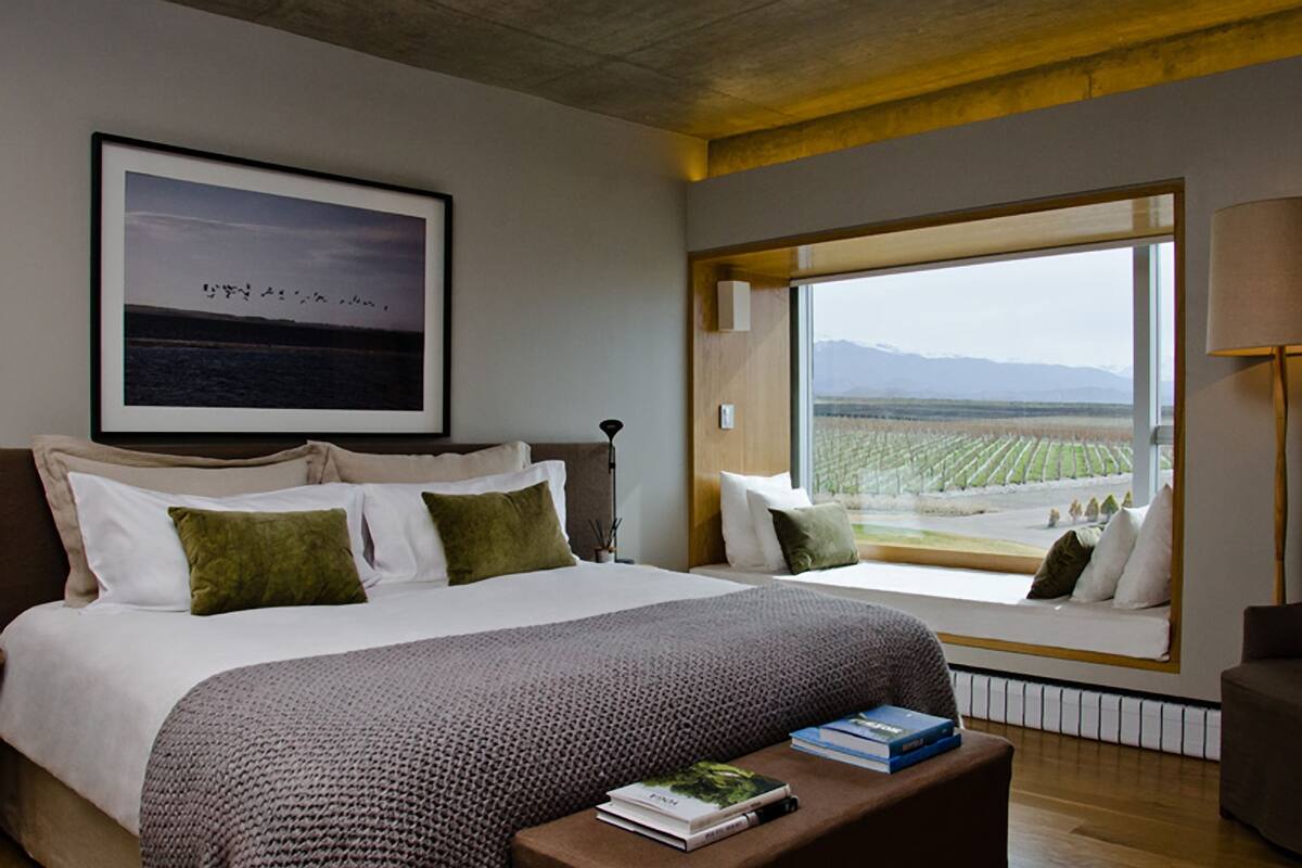 A bedroom with a large window, a bed with pillows, a bedside table with books, and a lamp.