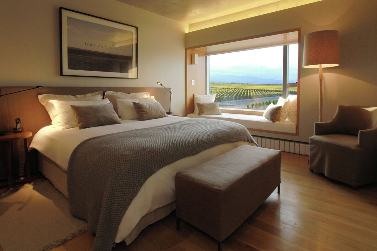 A bedroom with a large bed, a window with a view of a vineyard, a chair, and a bedside table.