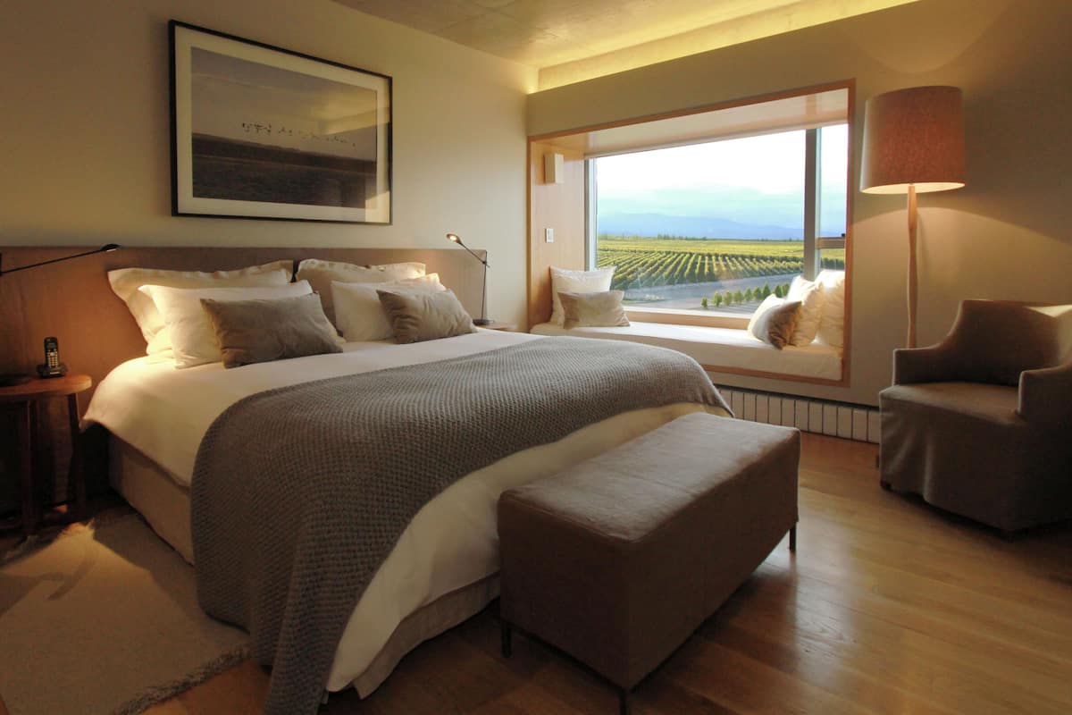 A bedroom with a large bed, a window with a view of a vineyard, a chair, and a bedside table.