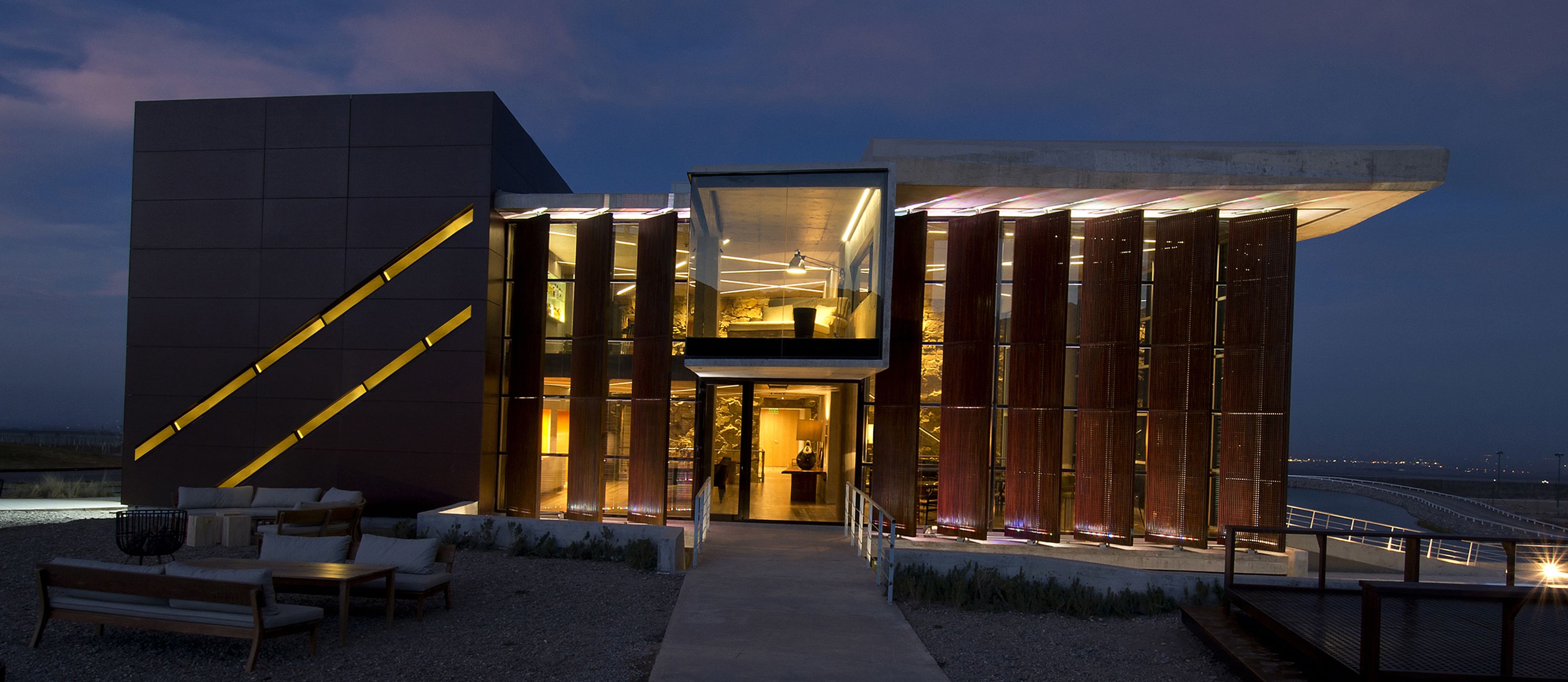 A modern building with a glass facade, a visible interior with a bed and a desk, and a pathway leading to the entrance.