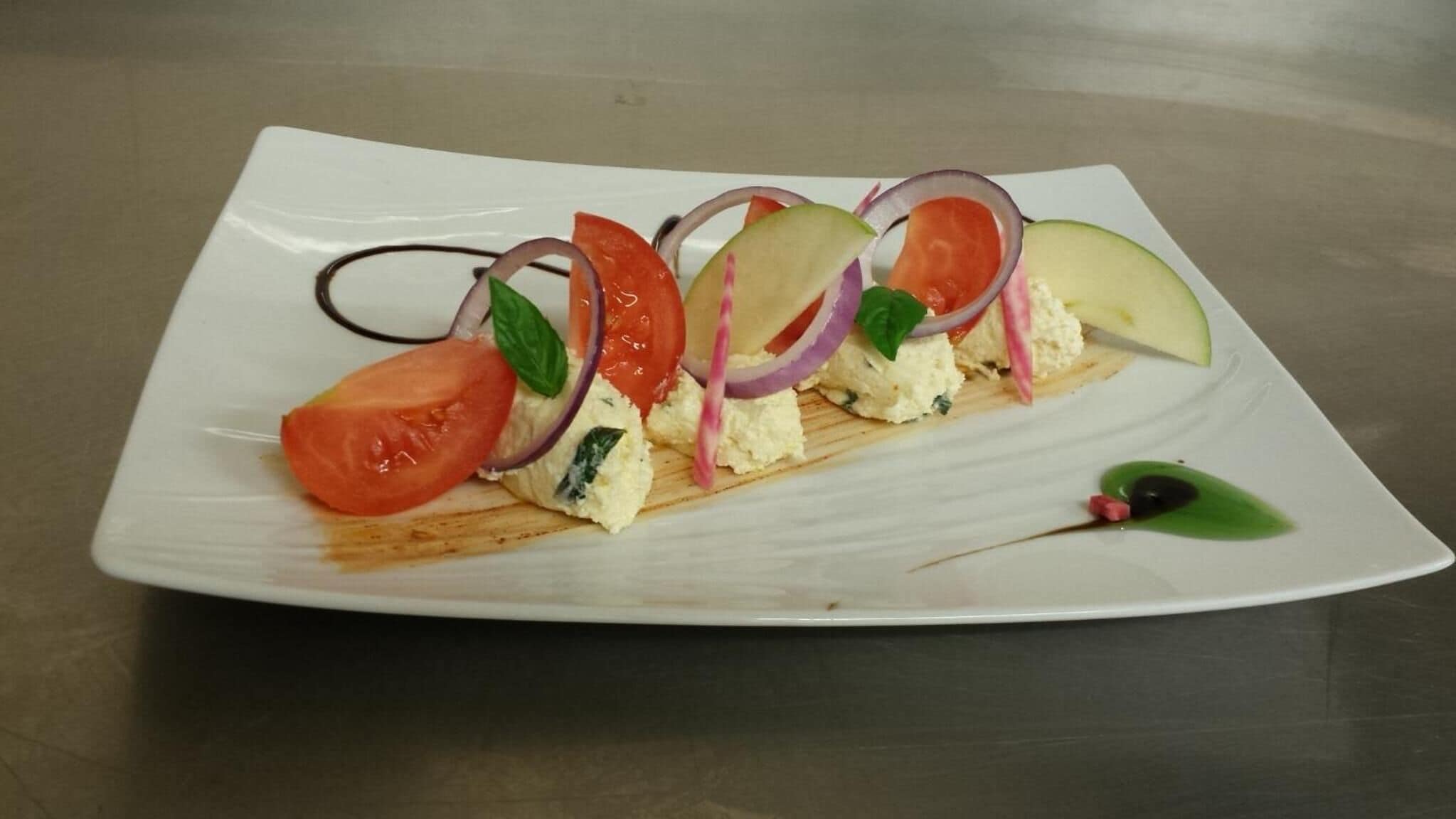 A white rectangular plate with a layered appetizer consisting of cheese, tomatoes, and onions.