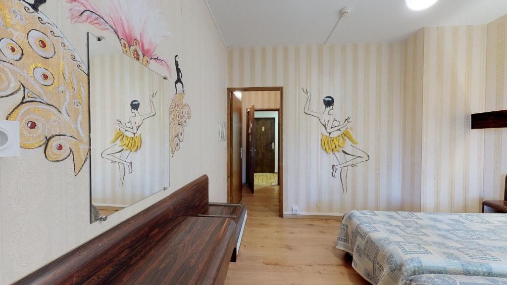 Quarto de hotel com um banco de madeira, cama com roupa de cama estampada e um espelho refletindo obras de arte na parede.