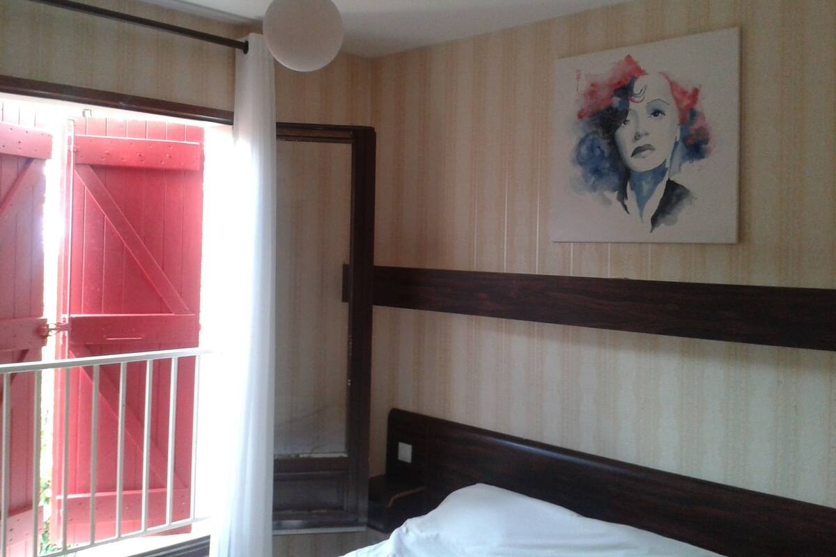 A hotel room with a bed, a painting on the wall, a window with a view of a red structure, and a staircase.