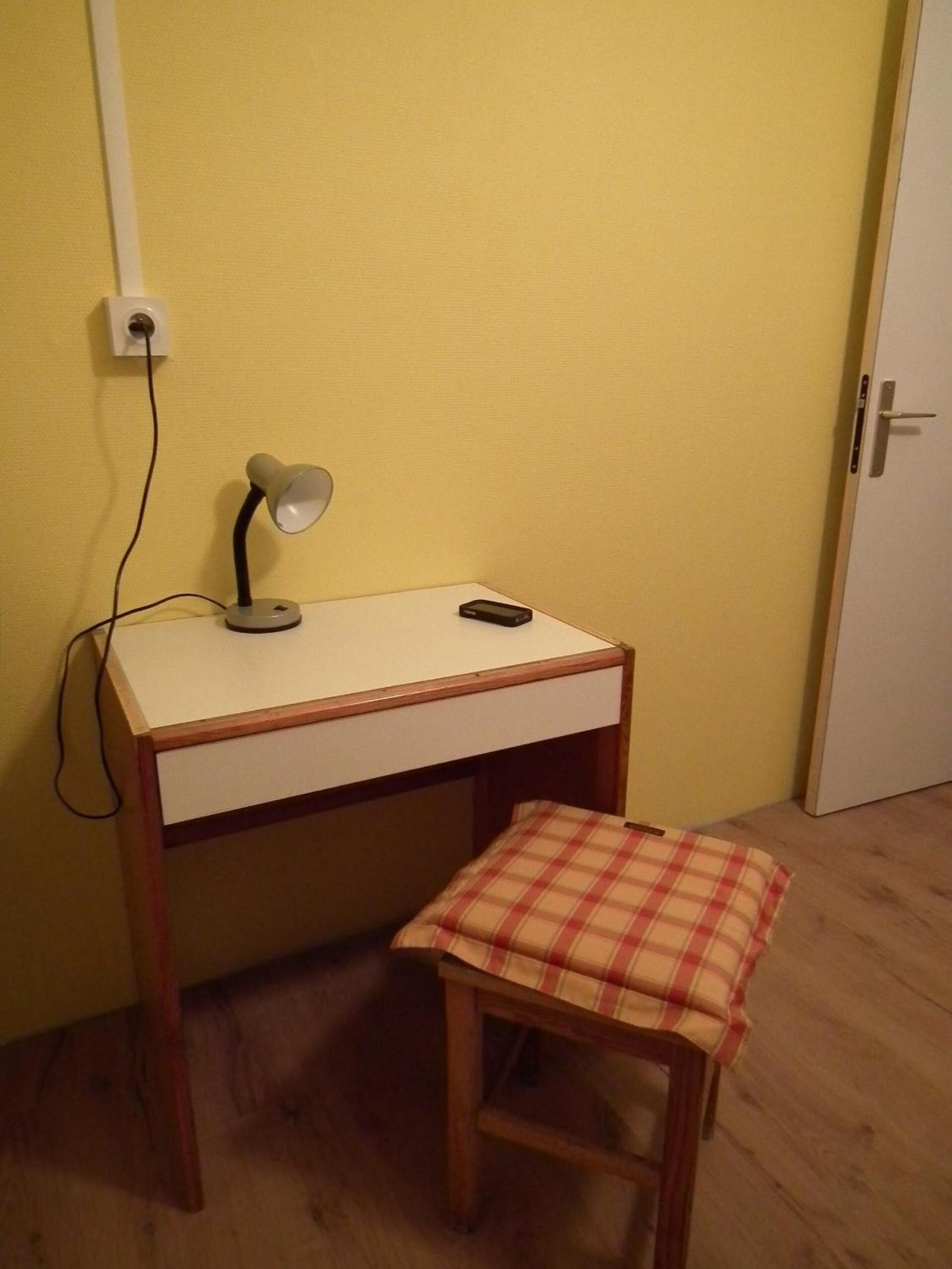 Pieni työpöytä, jossa on lamppu, matkapuhelin ja ruutukuvioinen koristetyyny puisen jakkaran päällä.