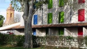 A building with a white facade, green and red shutters, and a tree in the foreground. - Friendly Auberge - Hostel (Colomiers)