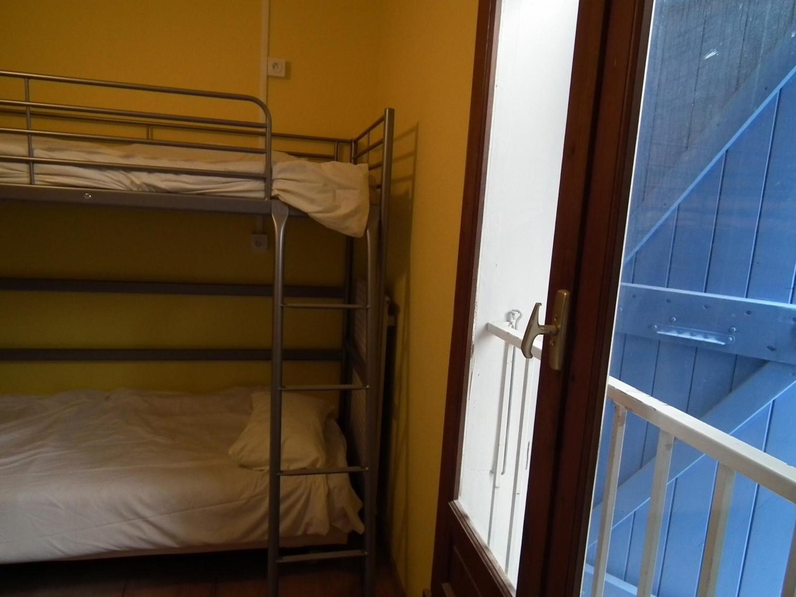 A dormitory room with bunk beds, a wooden door, and a view of a blue exterior.