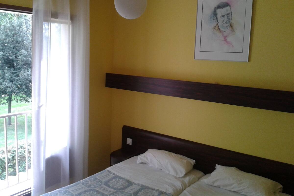 A bedroom with a bed, a window with curtains, a painting on the wall, and a ceiling light.