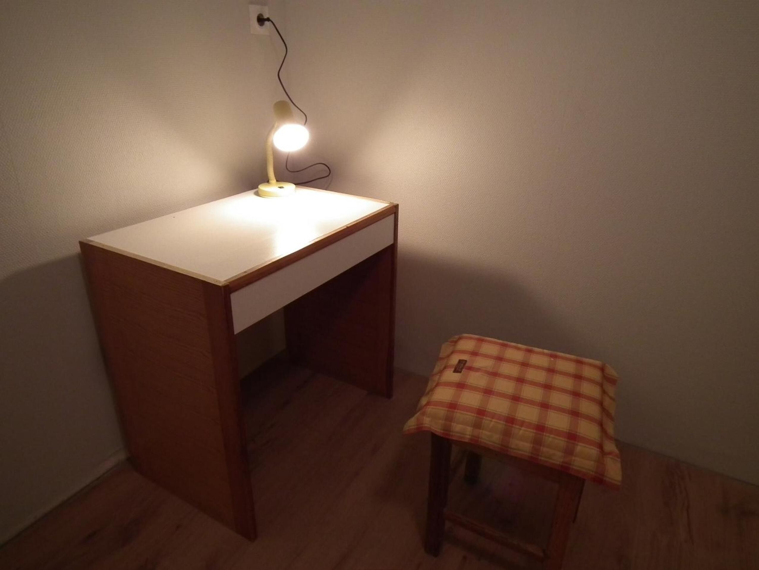 A small wooden desk with a white drawer and a lamp on top. There is a plaid tablecloth on a small table beside the desk.