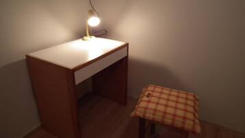 A small wooden desk with a white drawer and a lamp on top. There is a plaid tablecloth on a small table beside the desk.