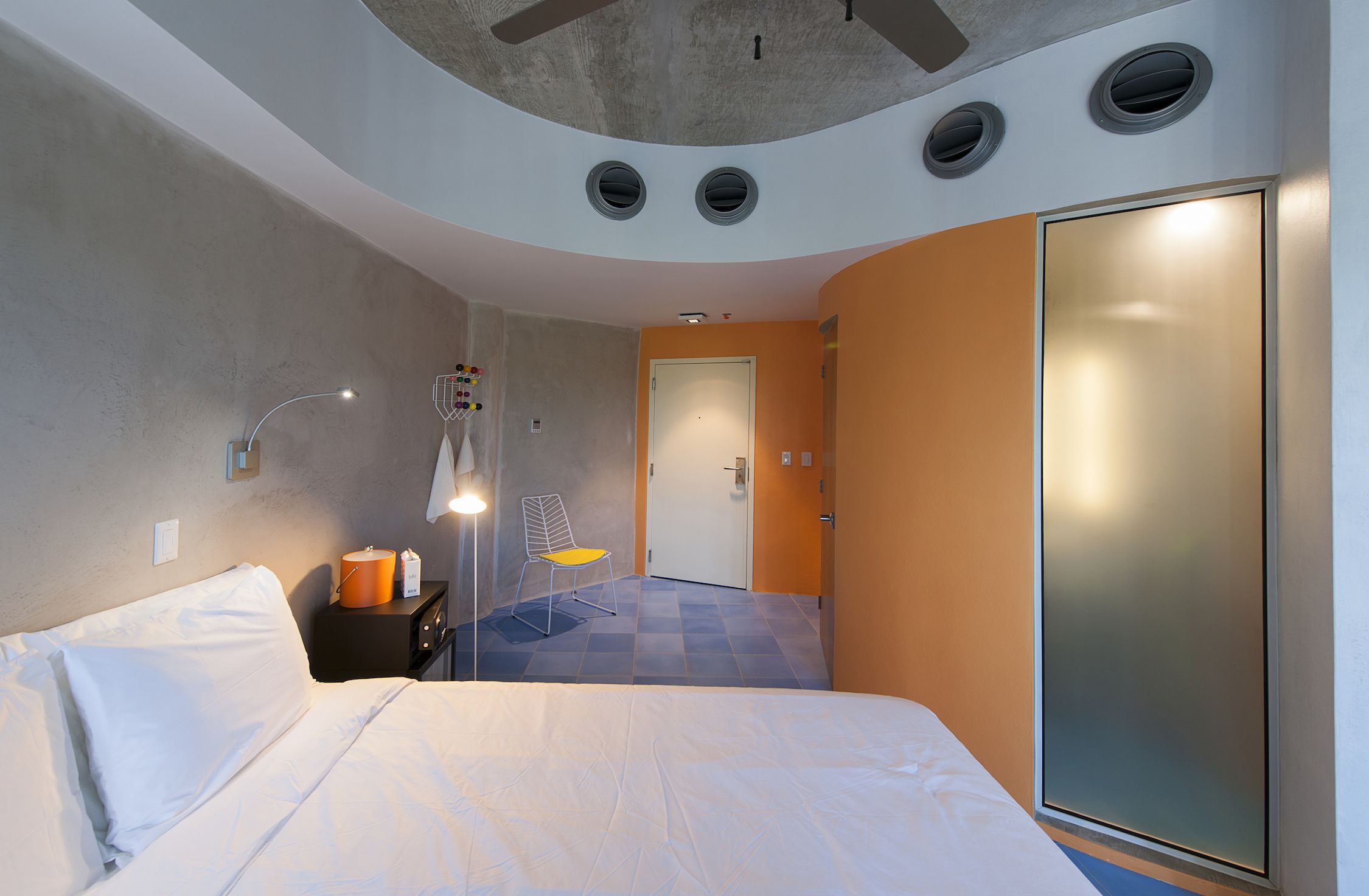 A modern hotel room with a large bed, a chair, a nightstand, a lamp, and a wardrobe.