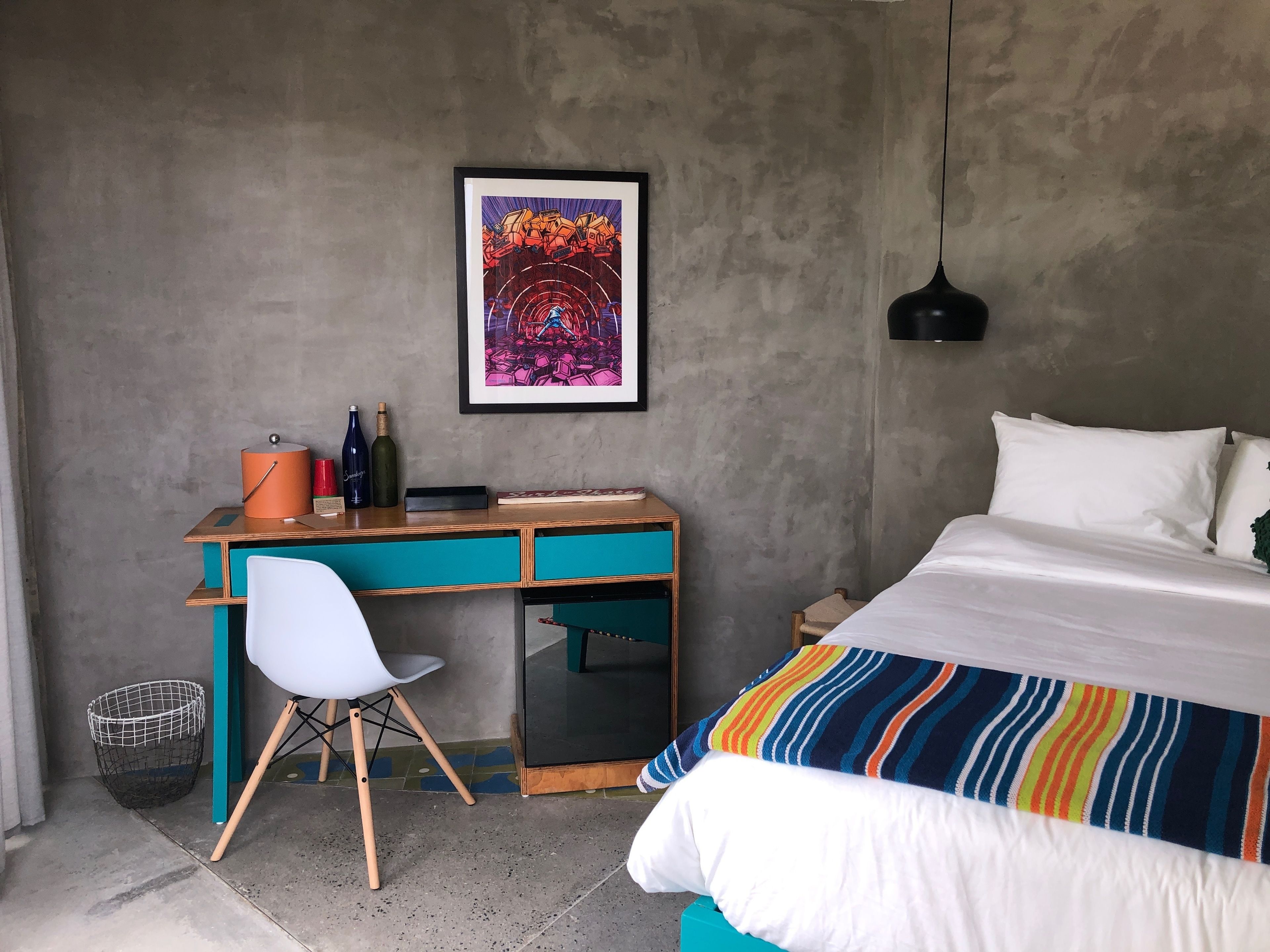 Un dormitorio con cama, un escritorio y una silla, un cuadro enmarcado en la pared y una lámpara colgante.