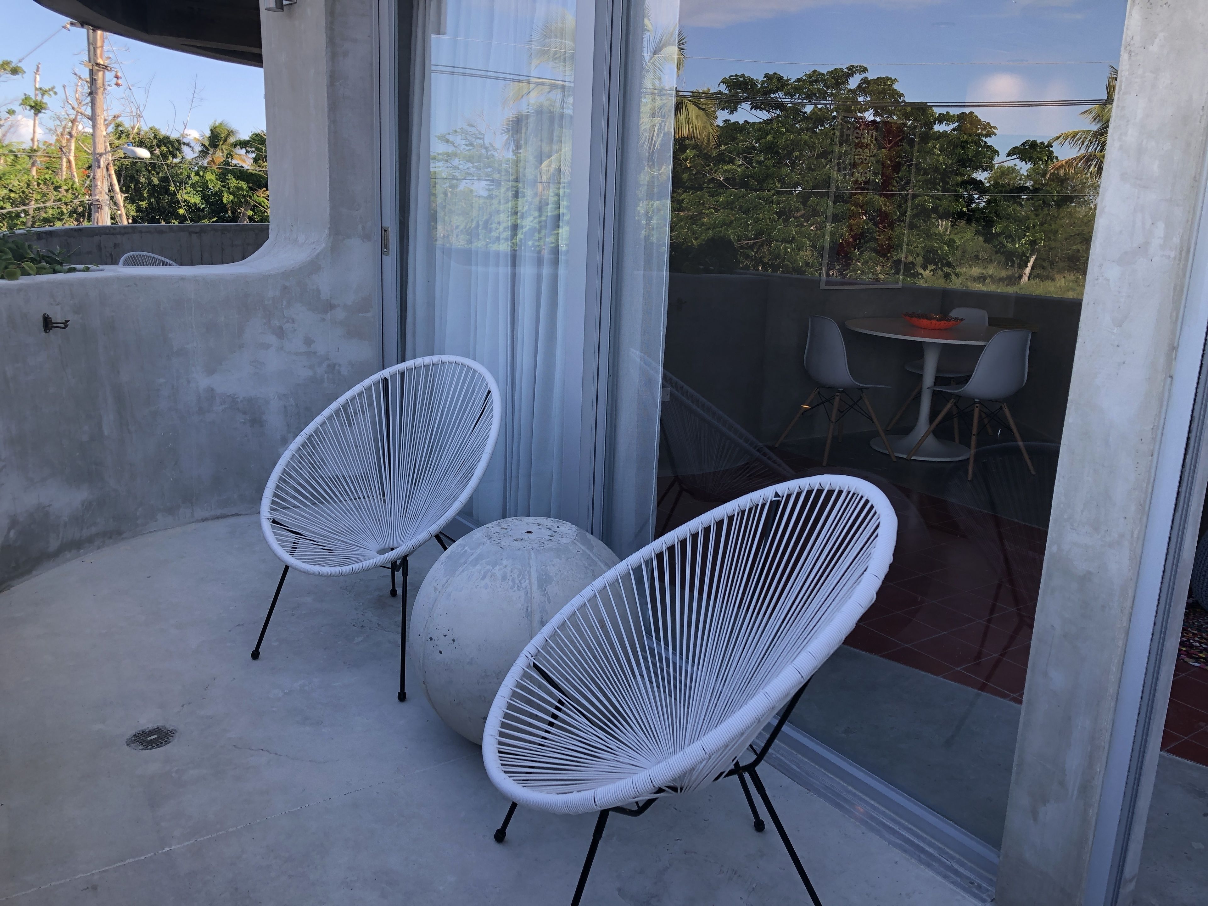 Un balcon avec deux fauteuils en osier blanc et une table ronde, donnant sur un jardin arboré et un ciel dégagé.