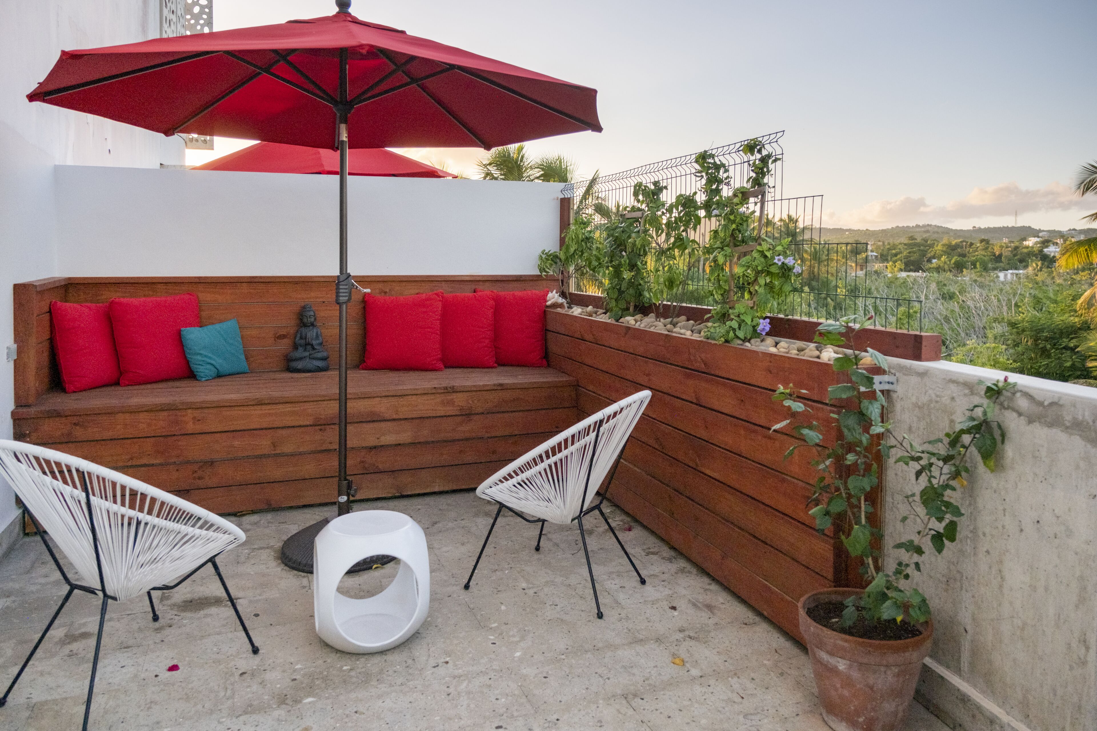 Un espace de détente aménagé sur le toit, avec un banc en bois, des coussins rouges, des chaises blanches et un parasol rouge.
