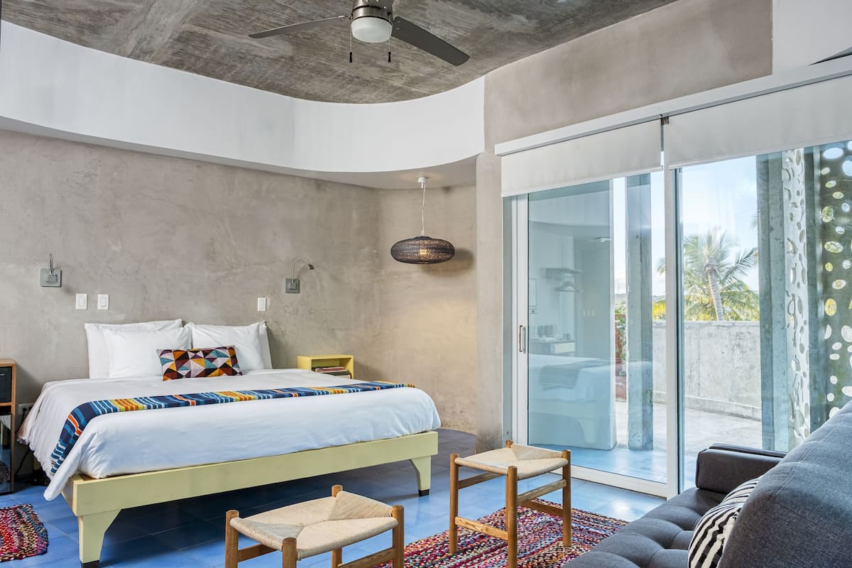 Un dormitorio moderno con cama, ventilador de techo, puerta de vidrio que da a un balcón y vistas a palmeras.