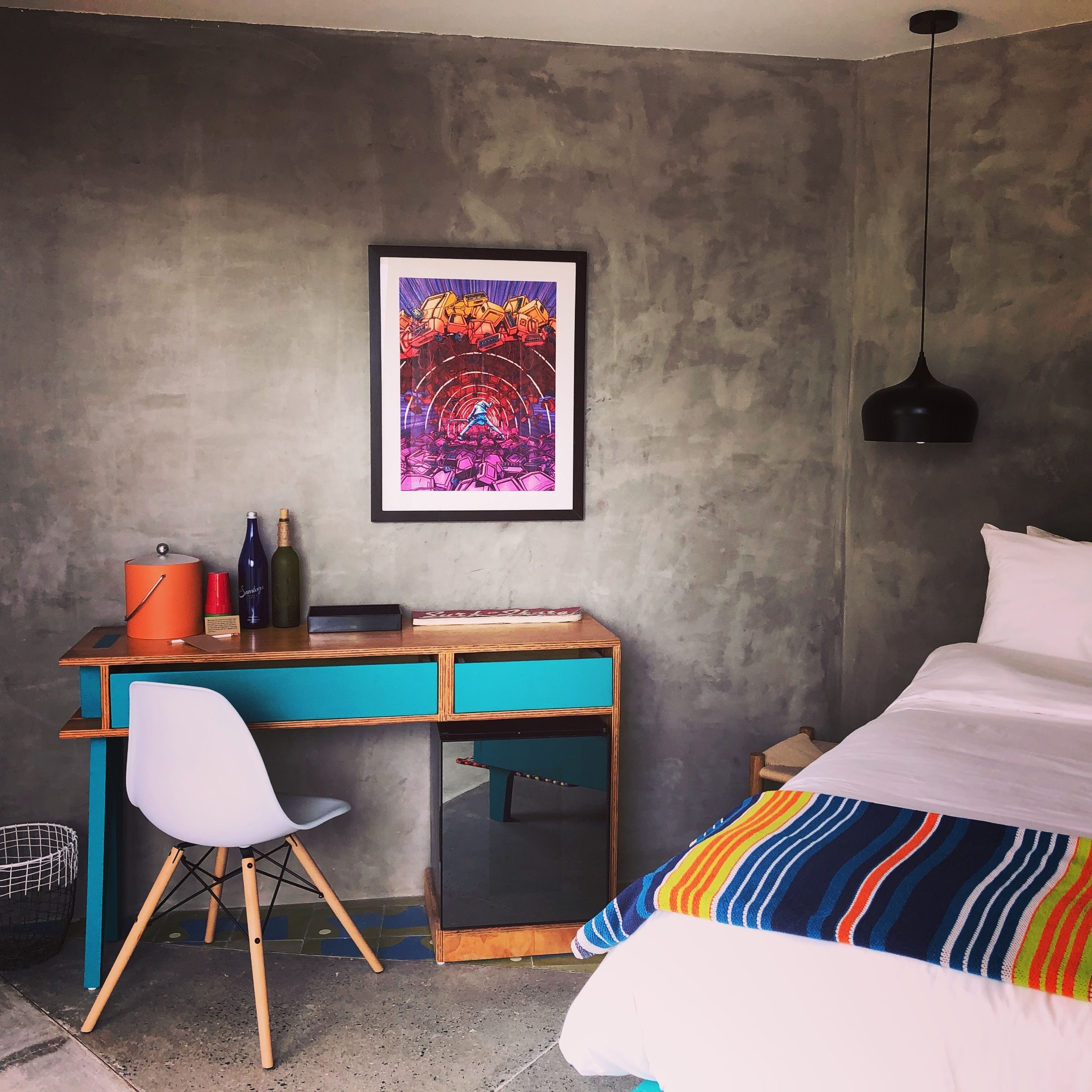 Une chambre à coucher avec un lit, un bureau avec une chaise, un tableau encadré au mur et un luminaire suspendu au plafond.