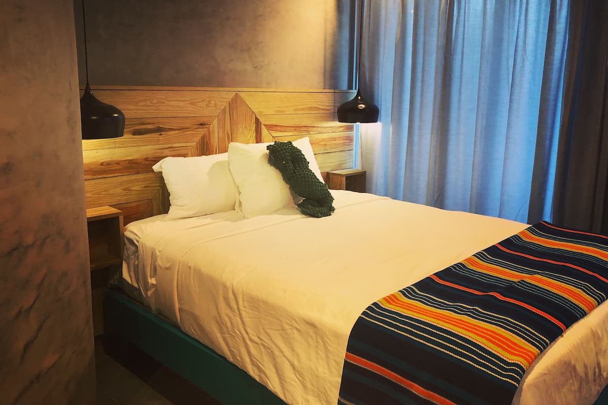 Una cama bien hecha con sábanas blancas y un colorido edredón a rayas, un cabecero de madera y una mesita de noche con una lámpara.