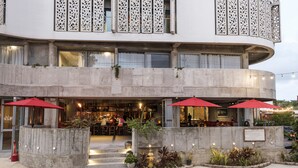 Un edificio de varios pisos con fachada curva, ventanas grandes y mesas al aire libre con sombrillas rojas.