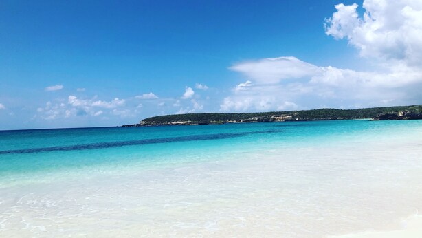 Una playa con agua turquesa cristalina, una orilla arenosa y una colina verde al fondo.