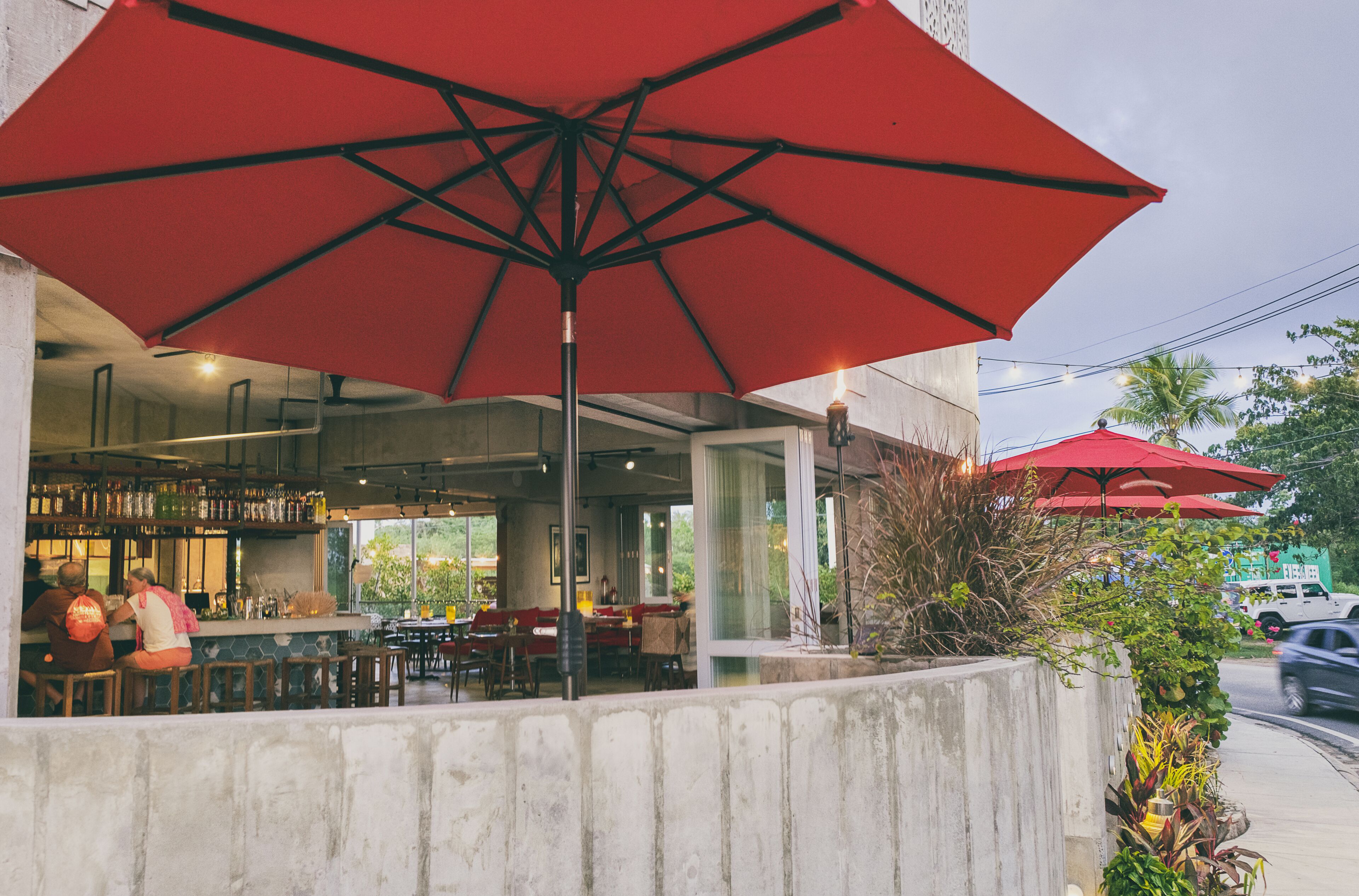 An outdoor seating area with red umbrellas, a bar, and a view of the street.