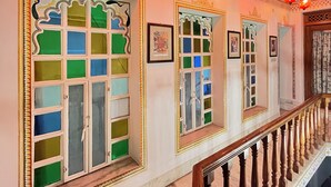 Ein Korridor mit aufwendiger Wandverzierungen, einer Treppe mit geschnitzten Holzbalkonen und mehreren Fenstern mit farbigen Glasfeldern.